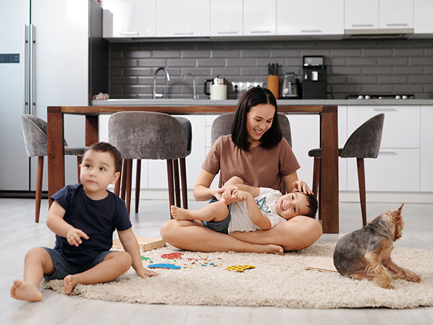 A mother plays with her two children and dog on the floor of their home.