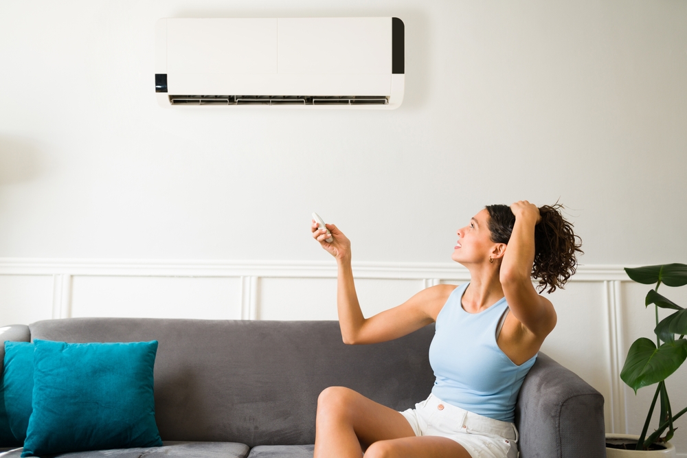Woman sitting on the couch with her hair pulled up in one hand while in the other hand she holds the remote to her ductless AC unit. She's hot and trying to get her AC to turn on.