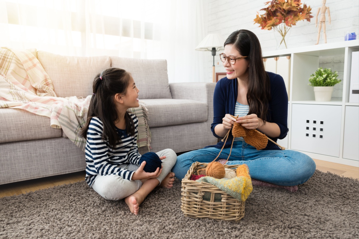 A young mother and daughter sitting on the floor as the mom knits and the girl tells her mom a story