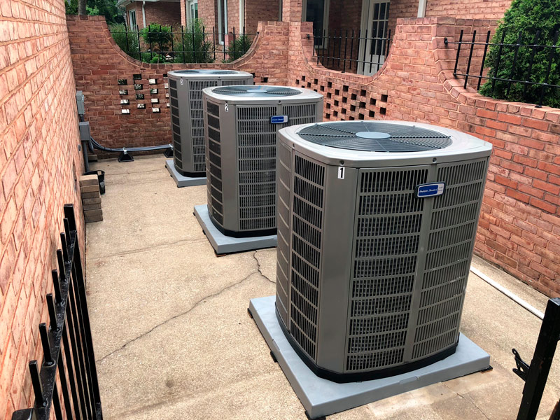 Multiple residential air conditioning units behind a brick building with a brick fence